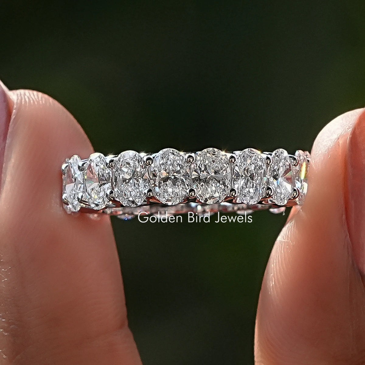Oval Cut Diamond Eternity Wedding Band Held Between Fingers