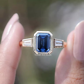 Front view of Blue Emerald Cut Moissanite Three Stone Ring in two fingers 