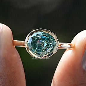 Front view of Blue Green Moissanite Step Cut Oval Solitaire Ring in two fingers 