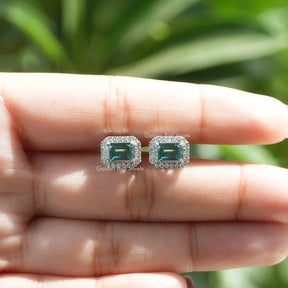 In a two-finger view of earrings in  green emerald cut moissanite