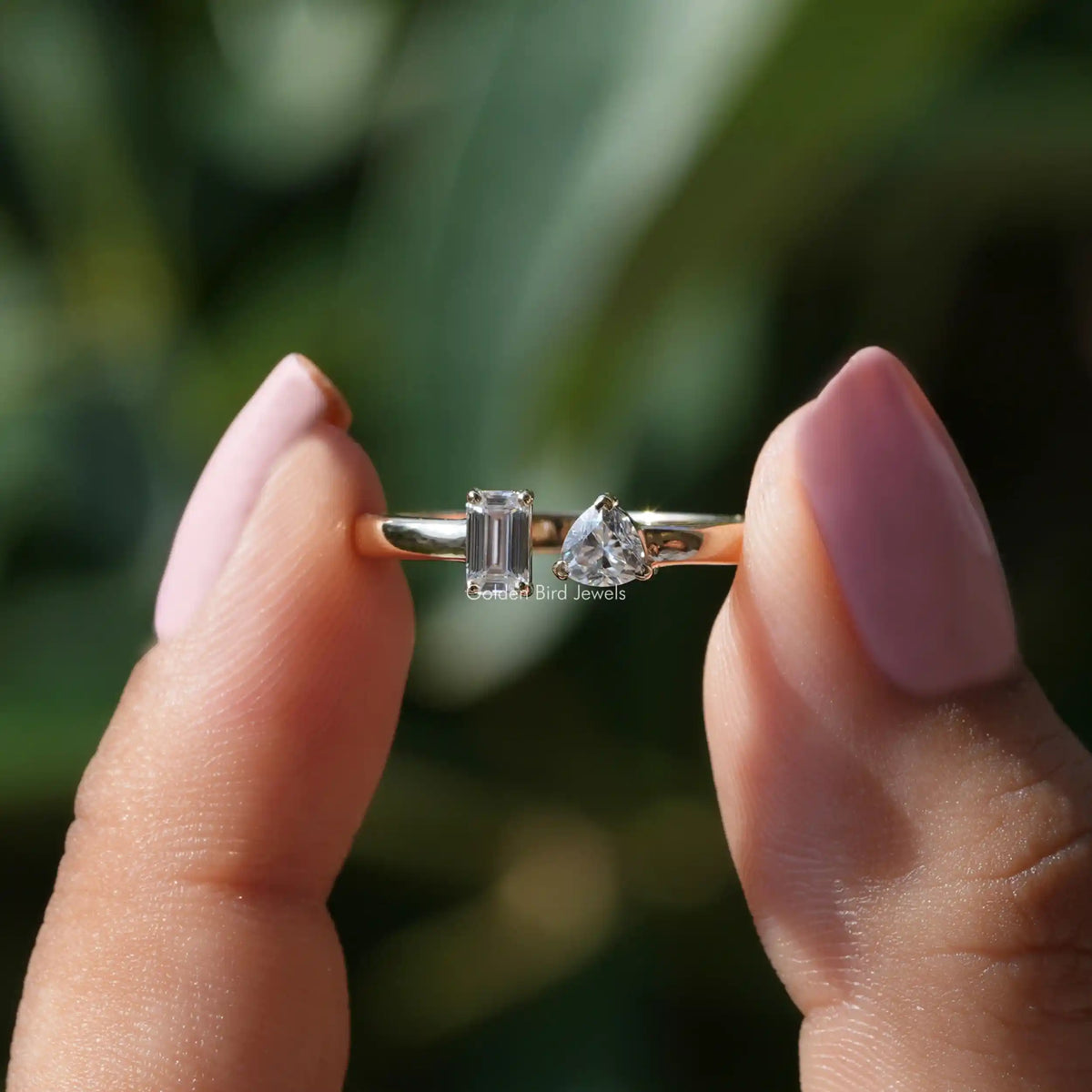 Two fingers holding the Emerald And Trillion Cut Moissanite Toi Et Moi Ring